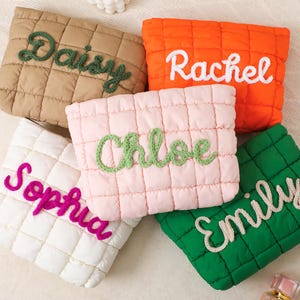 Puede incluir: Cinco bolsas acolchadas en varios colores: beige, naranja, rosa, blanco y verde. Cada bolsa presenta un nombre diferente bordado en un color contrastante: "Daisy", "Rachel", "Chloe", "Sophia" y "Emily".