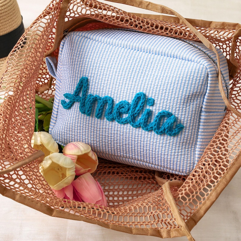 May include: A rectangular light blue and white seersucker pouch with a zipper closure, featuring the name Amelia embroidered in thick teal yarn on the front, placed inside a tan mesh tote bag alongside several artificial pink and cream tulips.