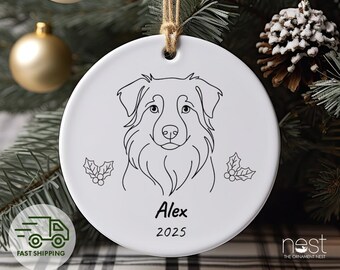May include: White ceramic Christmas ornament with a black line drawing of a dog's face. The ornament has the name "Alex" and the year "2025" printed on it. The ornament is hanging from a twine string.