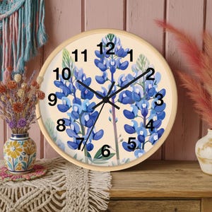 May include: A round wall clock with a light wood frame. The clock face features a cream background with bluebonnet floral illustrations and black hands. The numbers are in black. The clock is on a wooden surface.