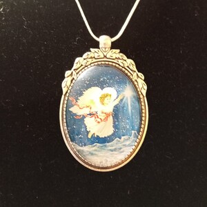 May include: A silver necklace with an oval pendant featuring a painted image of an angel with wings, flying through a snowy sky. The pendant has a decorative border of leaves.
