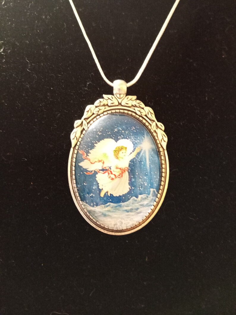 May include: A silver necklace with an oval pendant featuring a painted image of an angel with wings, flying through a snowy sky. The pendant has a decorative border of leaves.