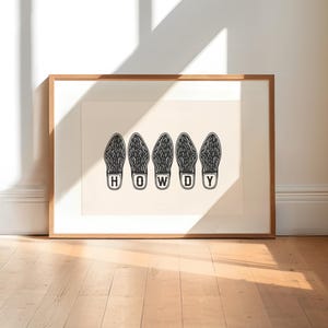 May include: A framed art print featuring five black and white shoe prints with the word "HOWDY" spelled out in white letters. The artwork is displayed on a light beige background, encased in a wooden frame.
