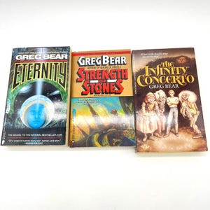 May include: Three science fiction novels by Greg Bear are displayed. The books are titled "Eternity", "Strength of Stones", and "The Infinity Concerto". The covers feature illustrations and text, with a green, yellow, and brown color scheme.