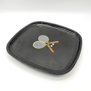 Puede incluir: Bandeja cuadrada negra con esquinas redondeadas. La bandeja presenta una ilustración dorada de dos raquetas de tenis cruzadas y dos pelotas de tenis blancas. Las raquetas tienen marcos dorados y mangos marrones.