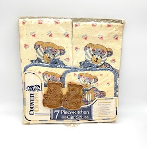 Puede incluir: Un juego de regalo de cocina de 7 piezas en un paquete de plástico transparente. El juego incluye dos paños de cocina beige y dos agarraderas, cada uno decorado con un diseño de oso de peluche. También se incluye una cuchara de madera y dos cortadores de galletas.