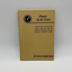 May include: A tan book cover featuring the title "Cheaper by the Dozen" in black script. The cover includes a black circular emblem and text detailing the dramatization and authors. The Dramatic Publishing Company is listed at the bottom.
