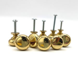 May include: A collection of six round, polished brass cabinet knobs, each featuring a smooth, reflective surface and a threaded metal screw extending from the back for installation. The knobs are arranged in a cluster, showcasing their gold-toned finish.