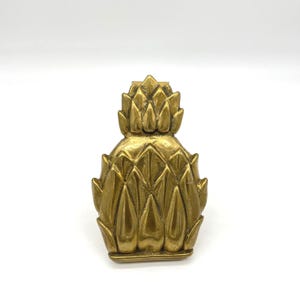 May include: A gold-coloured pineapple-shaped decorative object. The pineapple has a textured surface with detailed leaf and scale patterns. The object is likely made of metal and is designed to be a decorative accent piece.