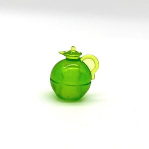 Puede incluir: Una jarra miniatura de vidrio verde lima translúcido con un cuerpo esférico redondeado, un pequeño pico vertedor y un asa curva. El objeto presenta un acabado liso y brillante con un tono verde claro y vibrante que permite el paso de la luz.