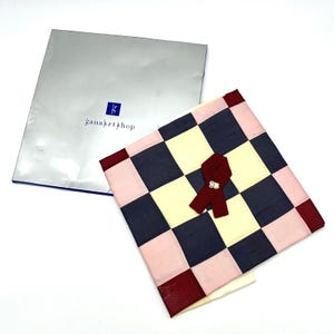 May include: A checkered pattern fabric in pink, navy, cream, and burgundy. A burgundy ribbon with a small decorative element is attached. A silver folder with the text "gana art shop" is in the background.