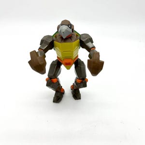 May include: A brown, yellow, and orange action figure of a turtle-like character. The figure has a bird-like head, a yellow shell, and articulated limbs. The figure is standing on a white surface.