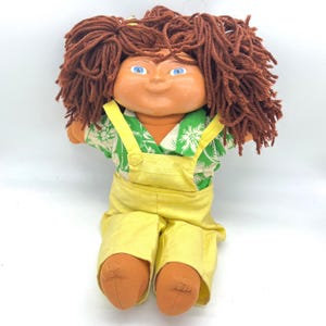 Puede incluir: Una muñeca vintage con cabello de hilo marrón, ojos azules y una cara bronceada. La muñeca lleva una camisa floral verde y blanca, un peto amarillo y zapatos color canela. Es un artículo de colección.