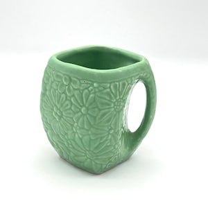 Puede incluir: Una taza de cerámica de color verde claro con un borde cuadrado y un cuerpo redondeado. La superficie exterior presenta un patrón floral en relieve que cubre todo el lateral, complementado con un asa curva a juego en el lado derecho.