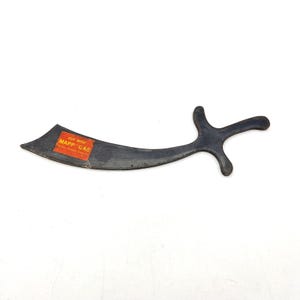 May include: A dark gray metal scimitar-shaped tool with a curved blade and a handle. A rectangular orange sticker is affixed to the blade, featuring the yellow text "CUT WITH MAPP GAS" and smaller text below it, indicating a promotional item.