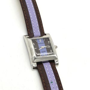 May include: A rectangular silver-tone watch with a black dial featuring a vertical lavender stripe and the text PARADE and QUARTZ. The watch has a brown leather strap with a central lavender inlay and white stitching along the edges.