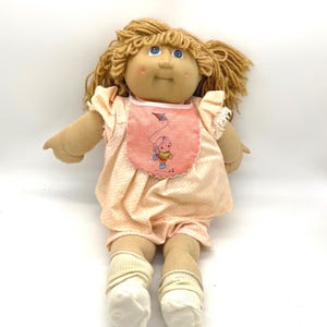 May include: A vintage doll with blonde yarn hair and blue eyes. The doll wears a peach-colored dress and shorts with white polka dots, a bib with a kite design, and white socks. The doll's skin is a light tan color.