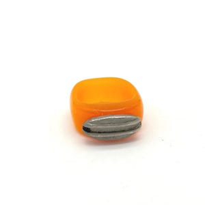 May include: An orange resin ring with a silver-colored, oval-shaped accent. The ring has a square shape with rounded corners. The silver accent is textured with horizontal lines. The ring is set against a white background.