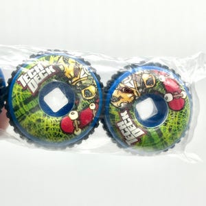 May include: Two Tech Deck skateboard wheels are visible, each with a blue rim and a central square hole. The wheels feature a green and yellow design with the words "TECH DECK" and a small red skateboard graphic.