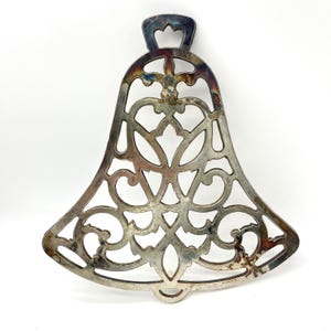May include: Silver-colored metal trivet in a bell shape with an openwork design. The trivet features a decorative pattern of swirling lines and heart-like shapes. It has a small loop at the bottom and a handle at the top.