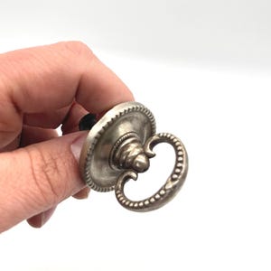 May include: A silver-toned metal drawer pull featuring a circular backplate with a beaded edge and a matching drop-style handle. The hardware has a weathered, metallic finish with a central mounting point and a curved, decorative loop design.