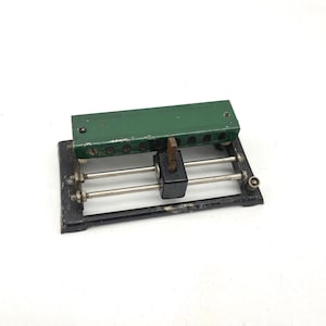 Puede incluir: Una herramienta antigua de estilo industrial con una parte superior verde y una base negra. La herramienta tiene una serie de agujeros a lo largo de la parte superior verde y varias varillas y tornillos metálicos. La herramienta parece ser de metal y probablemente se utiliza con fines mecánicos.