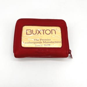 May include: A red leather Buxton wallet with a zipper closure. The wallet features a clear window and the text "BUXTON The Premier Leathergoods Manufacturer Since 1898".