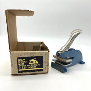 May include: An Ideal Model M1 R steel desk seal with a blue base and silver handle, next to its original cardboard box. The box has a black label with the text "STEEL DESK SEAL 1" X 2" INSERT".
