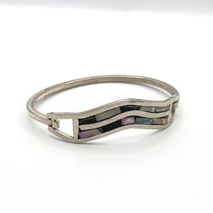 May include: Silver-tone hinged bangle bracelet with a wave-shaped inlay. The inlay features a mosaic of iridescent abalone shell pieces and black accents. The bracelet has a simple clasp closure.