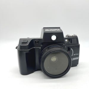 May include: Black Olympia L2000 camera with a lens and a flash. The camera has the word "Olympia" printed on the top. The camera has a self-timer function.