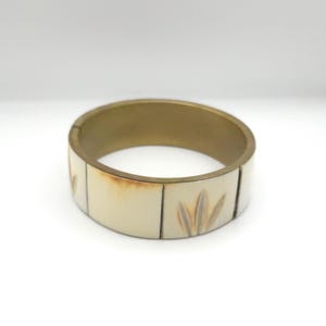 May include: A brass and ivory-colored bangle bracelet. The bracelet features a wide band with a gold-colored interior and ivory-colored exterior segments. Each segment has a leaf design. The bracelet is a piece of jewelry.