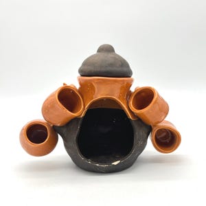 May include: A ceramic sculpture in shades of brown and orange, featuring a central structure with a dark brown lid and four attached cup-like elements. The piece has a rustic, handmade appearance, with a matte finish.
