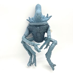 May include: A light blue action figure of a creature with a large head, detailed chest, and multiple limbs. The figure has a textured appearance and is posed in a seated position. The figure is on a white background.
