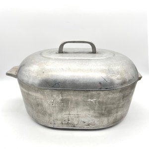 May include: An oval-shaped aluminium roasting pan with a matching lid. The metal surface features a brushed, matte finish with visible signs of wear, including light scratches and scuffs. It has two side handles and a central handle on the lid.