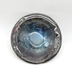 May include: A round ceramic bowl with an irregular, wavy rim. The interior features a glossy glaze with a blend of deep blue, metallic charcoal, and hints of iridescent purple. The surface has a textured, reflective finish with visible light highlights.