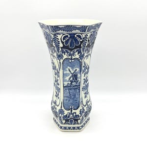 May include: A hexagonal ceramic vase featuring a cream-colored base with intricate blue floral patterns and a central panel depicting a windmill scene. The vase has a flared rim and a tapered body, showcasing detailed blue illustrations.