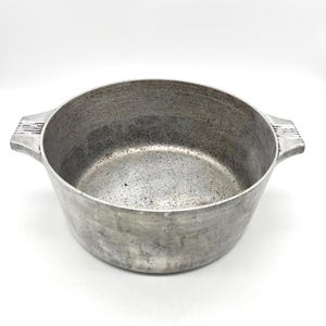 May include: A round, silver-coloured metal cooking pot with a textured, weathered finish. The pot features two integrated side handles, each displaying a series of horizontal grooved lines on the top surface. The interior shows signs of use.