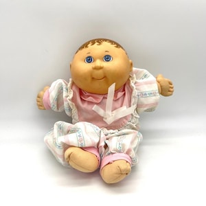 Puede incluir: Una muñeca de bebé vintage con tez bronceada, ojos azules y cabello castaño. La muñeca lleva un babero rosa y un conjunto a juego con un estampado floral y de rayas. Los brazos y las piernas de la muñeca están extendidos.
