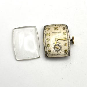 May include: A rectangular Bulova watch face with a silver dial, gold-toned numerals from 1 to 12, and gold hands. A small sub-dial is positioned at the bottom. The watch includes a separate, detached rectangular clear glass crystal cover.