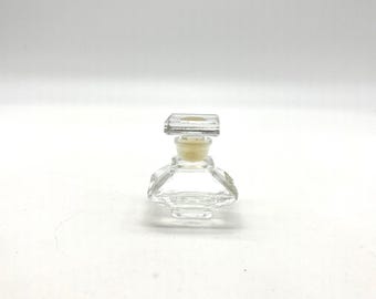 Puede incluir: Un pequeño frasco de perfume de vidrio transparente con un diseño geométrico. El frasco tiene un tapón cuadrado y un corcho beige. El cuerpo del frasco tiene una forma facetada, que refleja la luz. El diseño general es vintage y elegante.