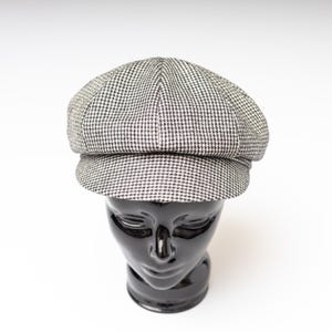 May include: A gray and white houndstooth pattern newsboy cap. The cap is displayed on a black mannequin head against a white background. The cap has a rounded crown and a stiff, flat brim.