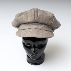 May include: A light brown newsboy cap featuring a subtle herringbone pattern. The hat has a structured, rounded crown with a short, stiff brim extending from the front, crafted from a woven fabric with a textured, multi-tonal appearance.
