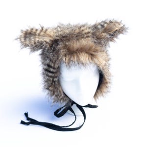 May include: A faux fur hat with ear flaps and a black tie closure. The hat features a mix of brown and tan fur, with darker stripes on the ear flaps. The hat is displayed on a white mannequin head against a white background.