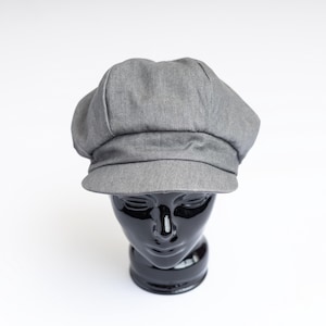 May include: A grey newsboy cap featuring a structured crown with multiple panels and a short, stiff brim. The hat is crafted from a textured fabric with a visible weave, displaying a classic rounded silhouette that sits atop a black mannequin head.