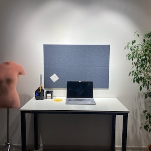 May include: A workspace featuring a white desk with a laptop, a dress form, and a blue felt board. A small plant and office supplies are also present. The dress form is a light orange color.