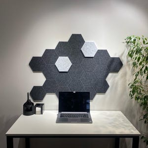 Könnte beinhalten: Ein modernes Büro mit einem weißen Schreibtisch, einem Laptop, einem schwarzen Dekorationsgegenstand und einer Ansammlung grauer und weißer sechseckiger Wandpaneele. Eine grüne Pflanze fügt dem Arbeitsbereich ein natürliches Element hinzu.