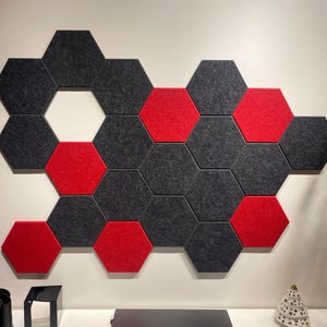 May include: Hexagonal acoustic wall panels in dark grey and red. The panels are arranged in a honeycomb pattern on a white wall. The design is suitable for sound dampening or decorative purposes, creating a geometric pattern.