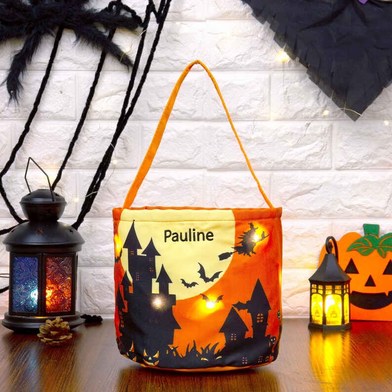 May include: A Halloween-themed trick-or-treat bag with an orange and black design. The bag features a spooky castle silhouette, bats, and a witch flying across a full moon. The name "Pauline" is printed on the bag.