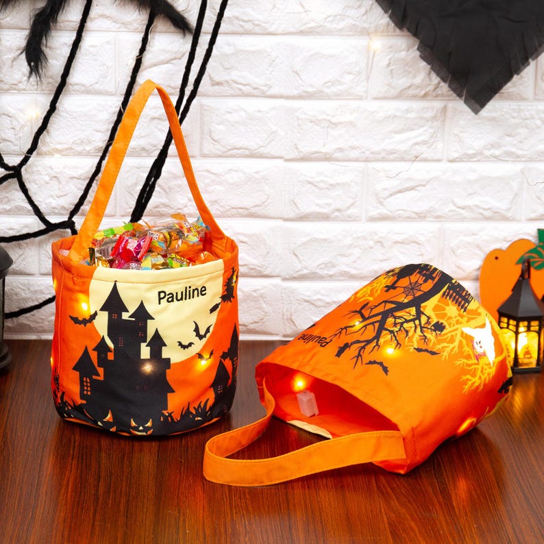 May include: Two orange Halloween treat bags with handles. One bag is upright and filled with sweets. The other bag is lying on its side. Both bags have a black castle and bat design, with the name "Pauline" printed on them.