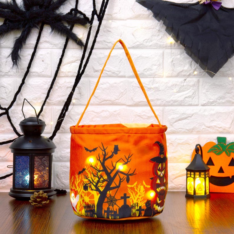 May include: Orange Halloween treat bag featuring a black tree, bats, and graveyard design. The bag has a handle and is illuminated with lights. A black spider and web are in the background, with a decorative lantern.